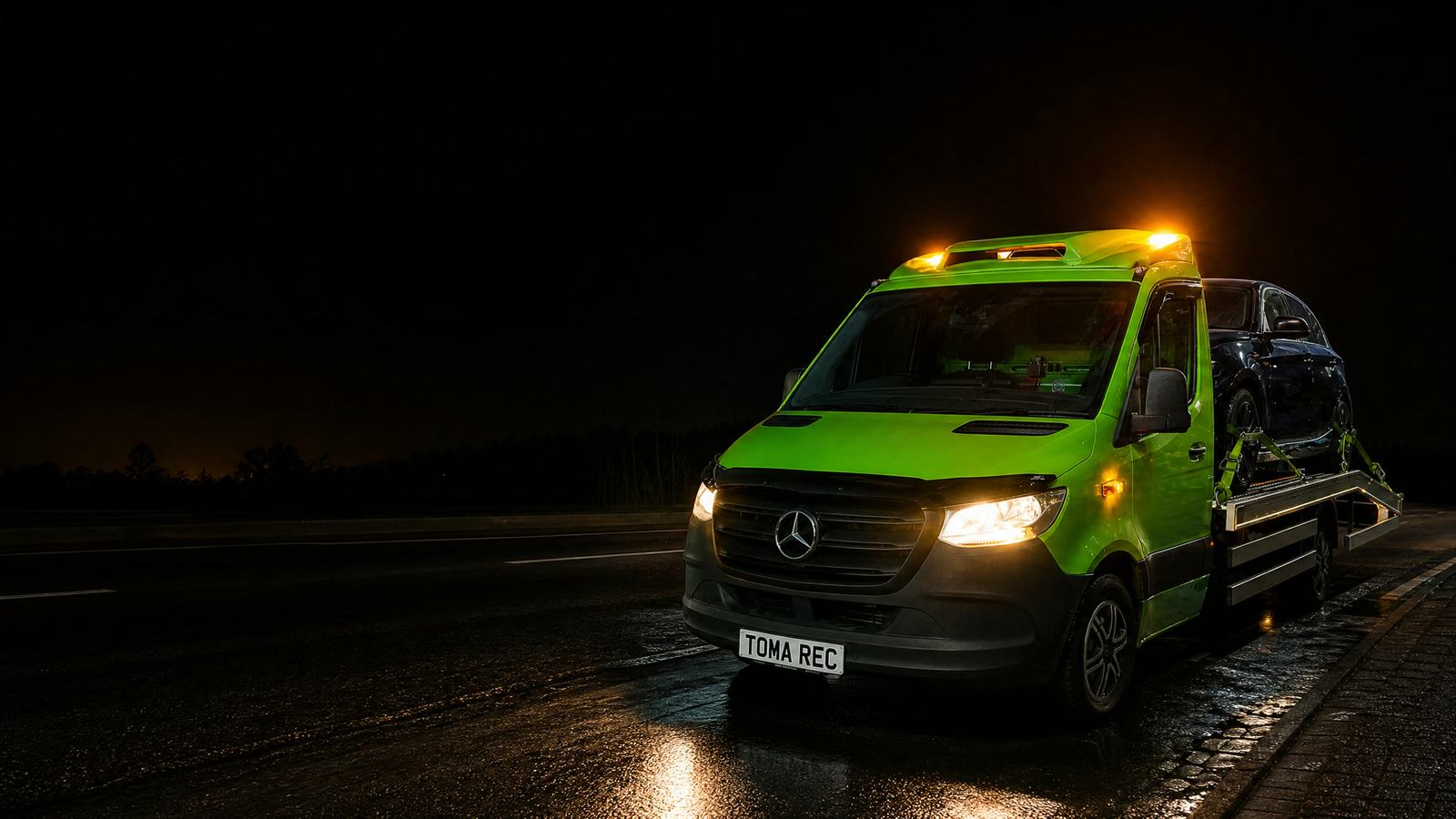 Toma Recovery's lime green Mercedes-Benz Sprinter flatbed at night on a wet lay-by, amber beacons on, a car strapped on the deck