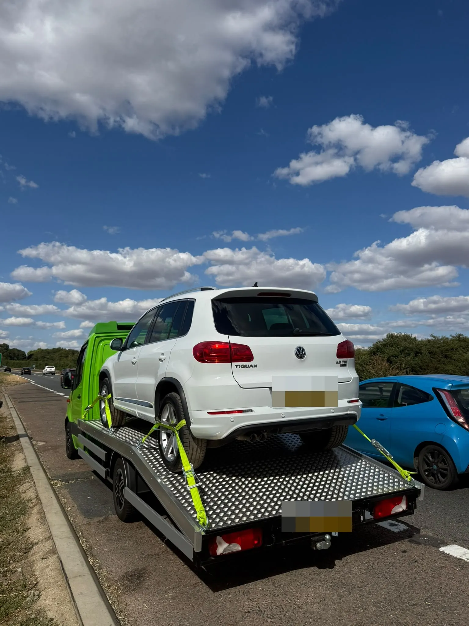 A white SUV strapped on Toma Recovery's flatbed parked on a grass verge beside a main road